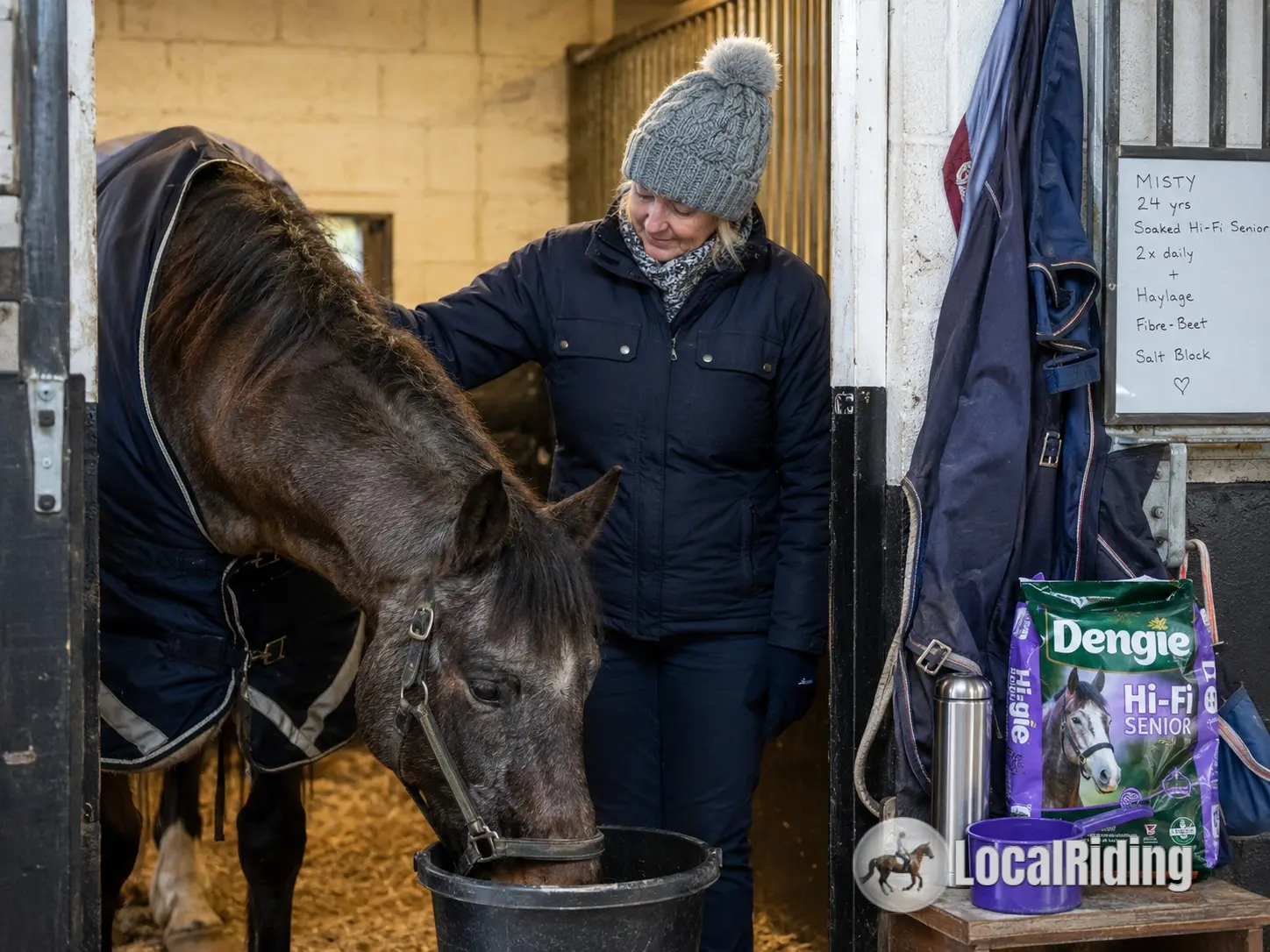 Owner feeding a soaked senior bucket feed to a rugged veteran horse in a stable in winter
